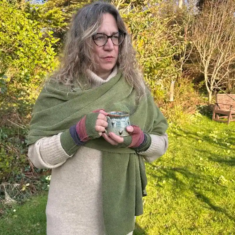Feeline Clothing green scarf/wrap worn with multi-coloured stripey mittens, pink, soft purple and green, by a woman holding a mug in a garden setting