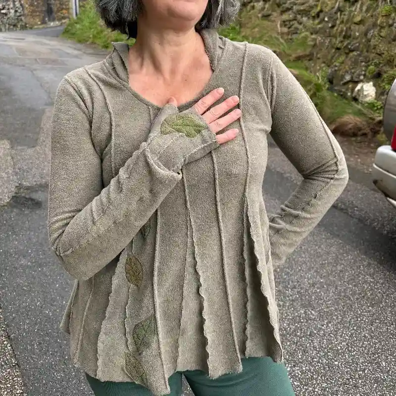 A Feeline Clothing top, with hood and long sleeves, in a soft warm neutral colour. There are leaves in shades of green appliquéd as an attractive design, front and back. Matching the top a pair of mittens are worn, in the same colours, also with leaves.