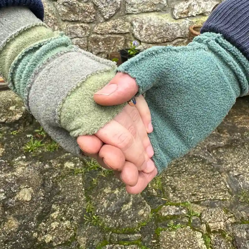 two hands holding each other wearing mittens in soft aqua and greens against an old stone wall