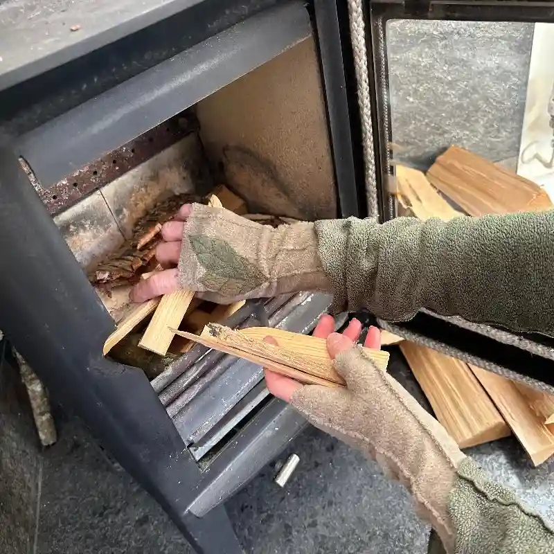 hands wearing beige mittens with a leaf appliqué design, putting kindling into a wood burner