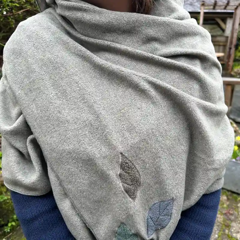 close up of a soft sage coloured fine fleece wrap/scarf, with blue and grey appliquéd leaves in one corner, worn over a blue jumper