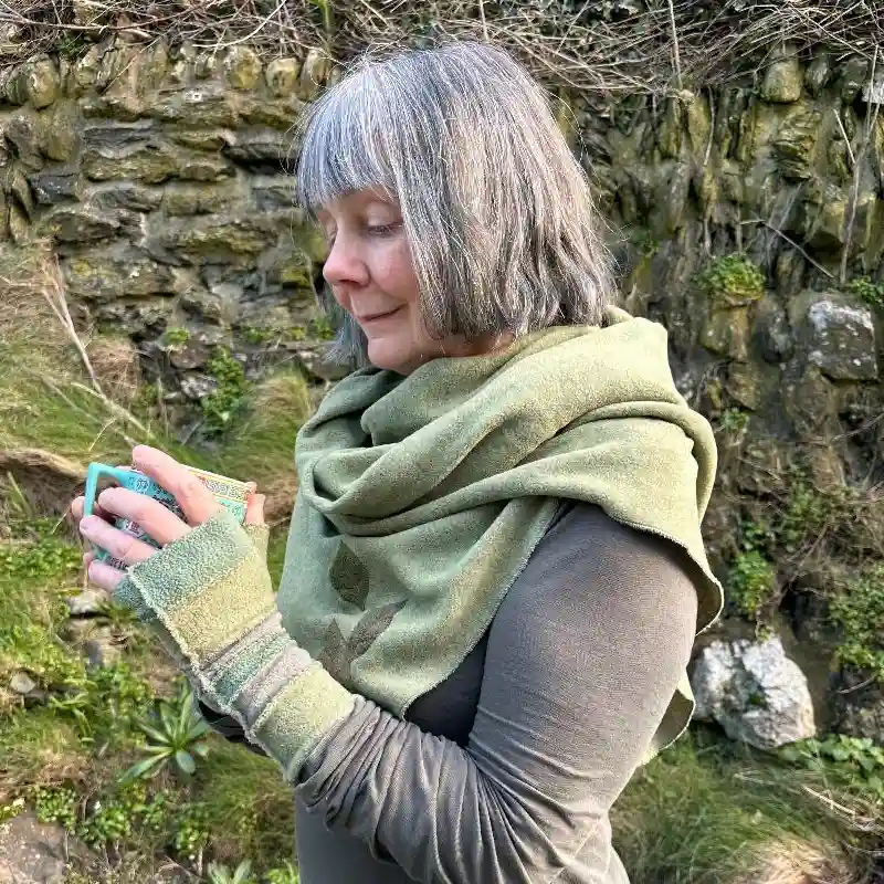 female mosel wearing a Feeline Clothing scarf/wrap and a pair of stripey mittens in soft shades of green, against a stone wall