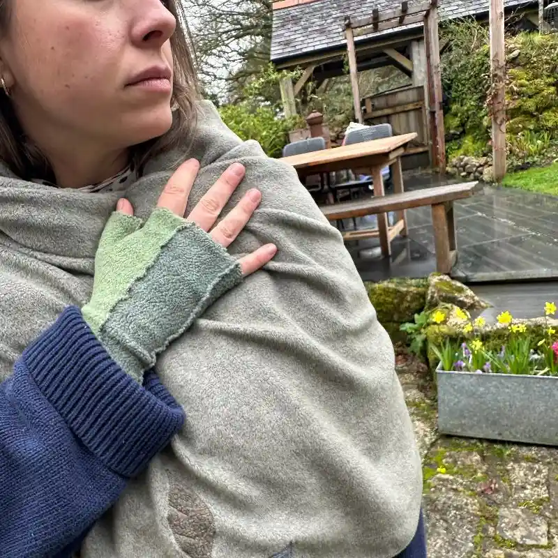 a model wearing a Feeline Clothing scarf/shawl and a two tone green mitten, in a garden setting